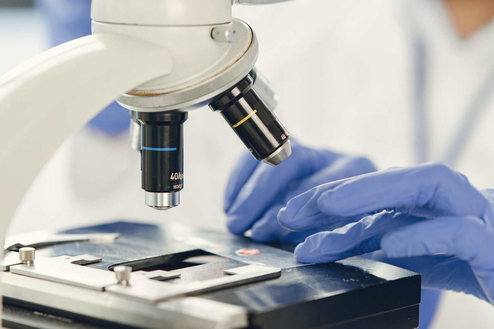 Gloved hands adjusting a specimen slide beneath the objective lenses of a laboratory microscope
