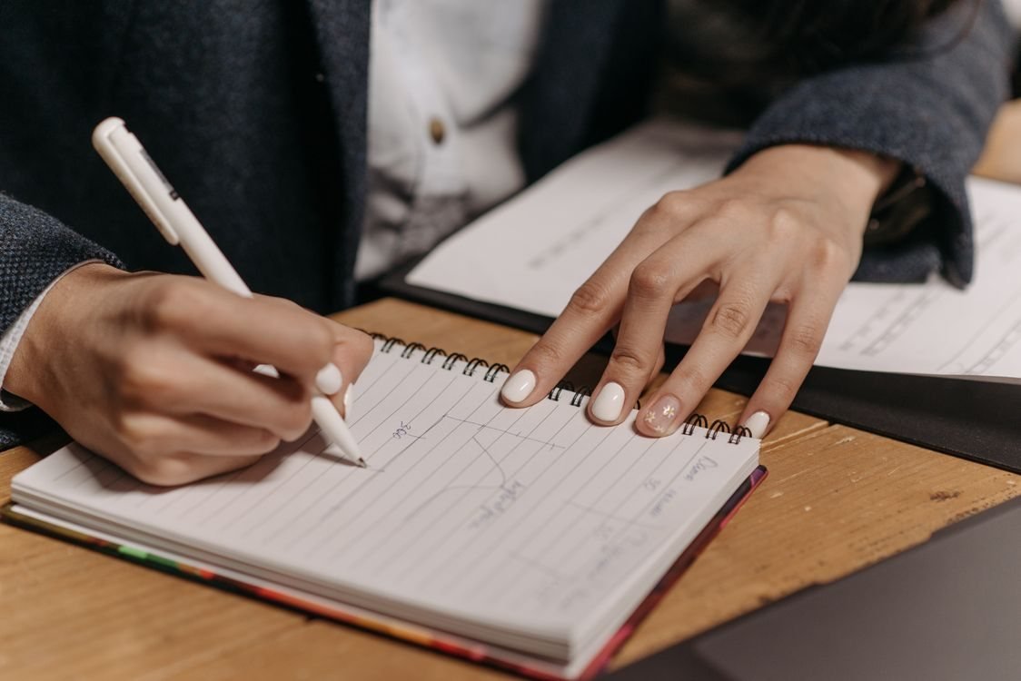 Close view of a person's hands writing figures and notes in a spiral-bound lab notebook on a wooden desk